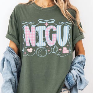 May include: A moss green t-shirt with a graphic of the word "NICU" in pink and blue checkered and striped patterns. The graphic is surrounded by hearts, stars, and blocks.