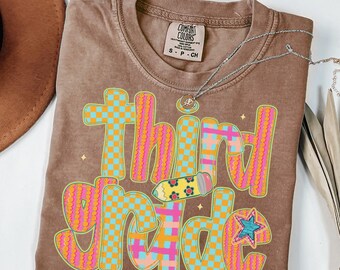 May include: Espresso-colored t-shirt with the words "Third Grade" in a colorful, checkered design. The design includes a pencil graphic and a star. The shirt is made of a soft material and has a round neckline. A silver necklace is draped across the shirt.