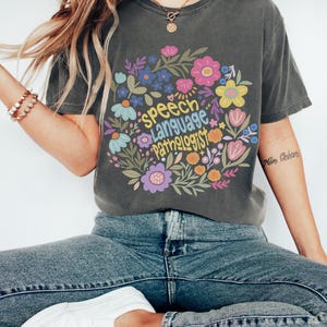 Puede incluir: Camiseta gris oscuro con un dise&ntilde;o floral y las palabras "Speech Language Pathologist" dispuestas en c&iacute;rculo. El dise&ntilde;o incluye flores y hojas coloridas. La camiseta es de corte c&oacute;modo.