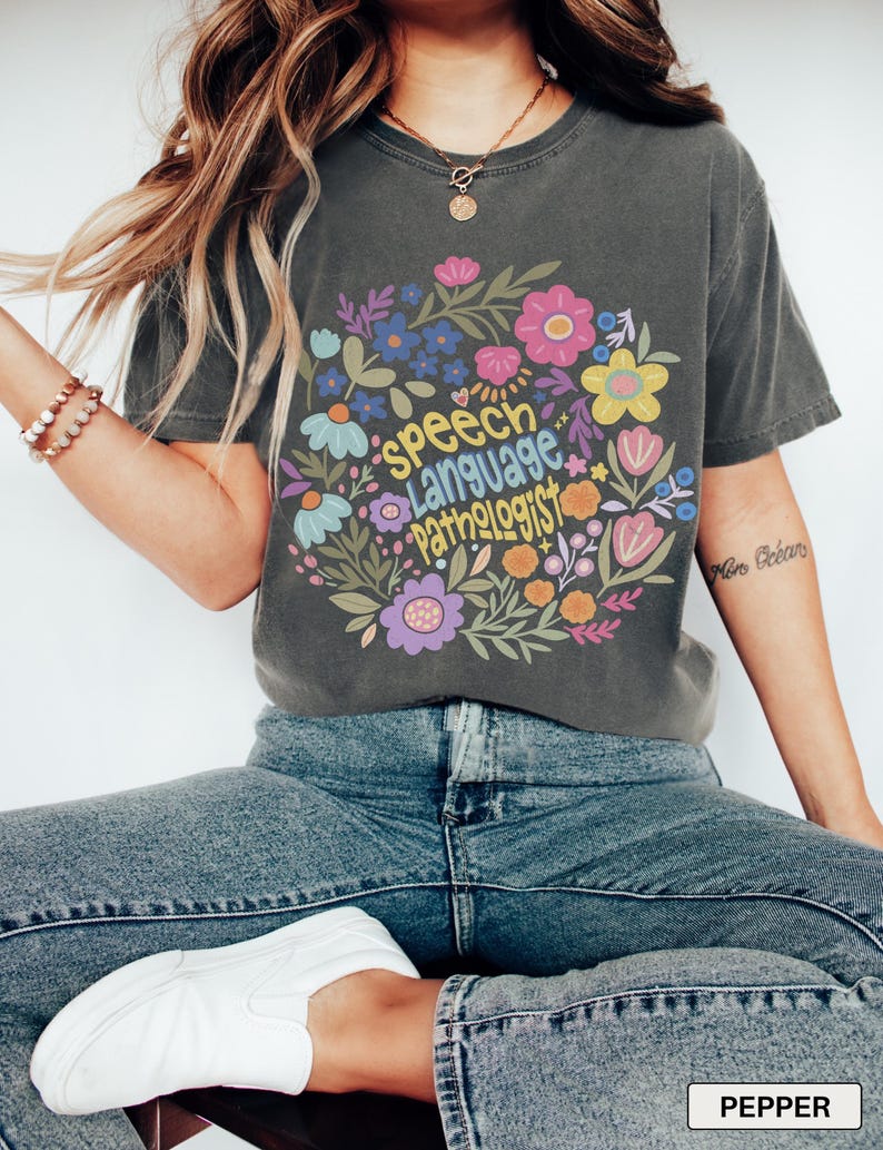 Puede incluir: Camiseta gris oscuro con un dise&ntilde;o floral y las palabras "Speech Language Pathologist" dispuestas en c&iacute;rculo. El dise&ntilde;o incluye flores y hojas coloridas. La camiseta es de corte c&oacute;modo.