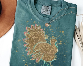 May include: Blue spruce Comfort Colors t-shirt with a turkey graphic. The turkey is brown with pink and teal accents, and gold speckles. A gold necklace with a pendant is on the shirt.