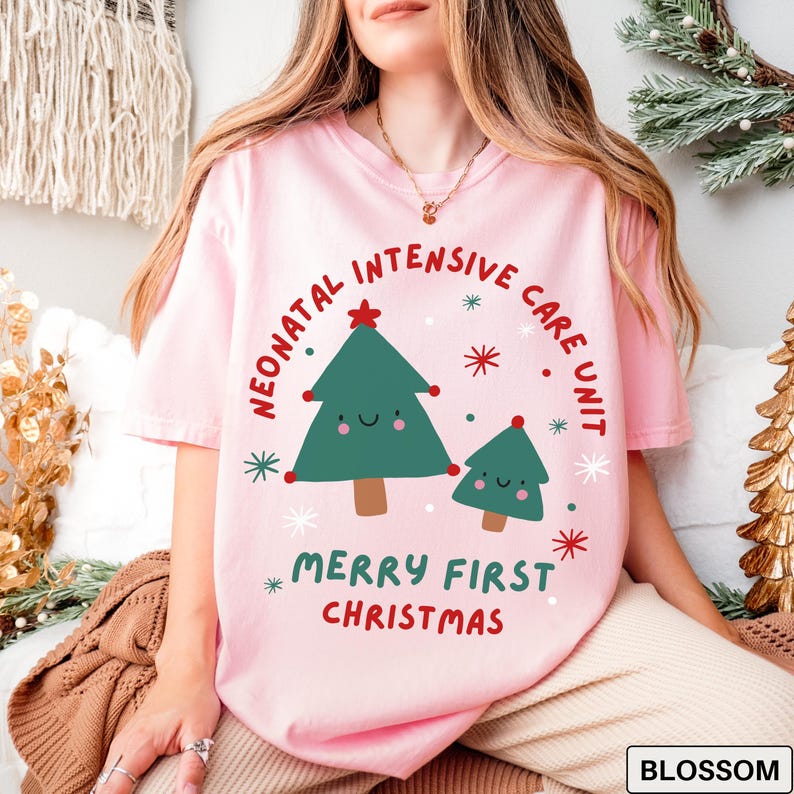 Comfort Colors blossom tshirt featuring a festive NICU design. The graphic shows two cute Christmas trees, one large and one small, surrounded by colorful red and green starbursts and snowflakes. Bold curved text reads “NEONATAL INTENSIVE CARE UNIT” above, with “MERRY FIRST CHRISTMAS” below in playful red and green lettering, creating a cheerful holiday look for NICU staff and families.