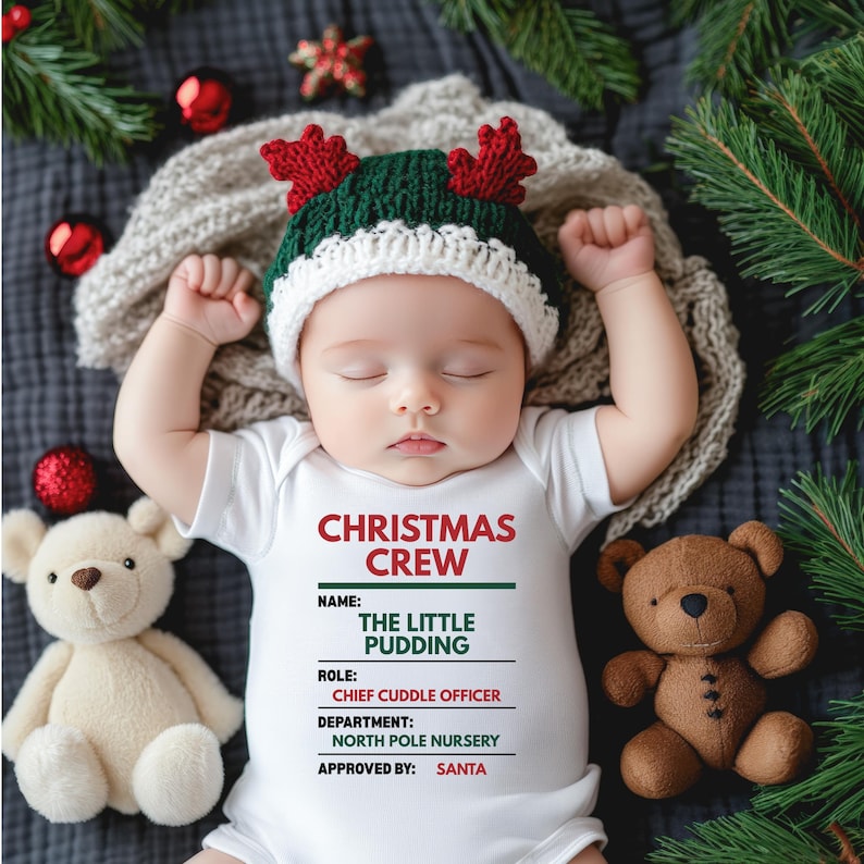 May include: A white baby bodysuit with red and green text reading CHRISTMAS CREW, NAME: THE LITTLE PUDDING, ROLE: CHIEF CUDDLE OFFICER, DEPARTMENT: NORTH POLE NURSERY, APPROVED BY: SANTA, worn with a green and white knit hat featuring red antlers.