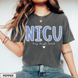 May include: Dark gray t-shirt with the word "NICU" in blue, dotted letters. The text "tiny, tough, loved" and a white heart are below the word. The shirt is a comfortable, casual style.
