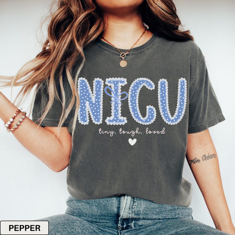 May include: Dark gray t-shirt with the word "NICU" in blue, dotted letters. The text "tiny, tough, loved" and a white heart are below the word. The shirt is a comfortable, casual style.