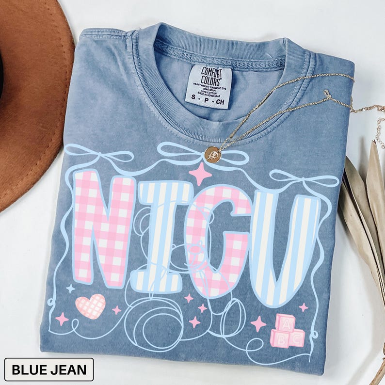 A folded blue jean Comfort Colors T-shirt with "NICU" spelled out in large, patchwork-style letters. The "N" and "C" are soft pink gingham, while the "I" and "U" are light blue and white vertical stripes. Surrounding the letters are pastel pink ribbons, looping thread, sparkles, a pink gingham heart, and pink ABC building blocks. The style is sweet, nurturing, and whimsical, perfect for NICU nurses and staff.
