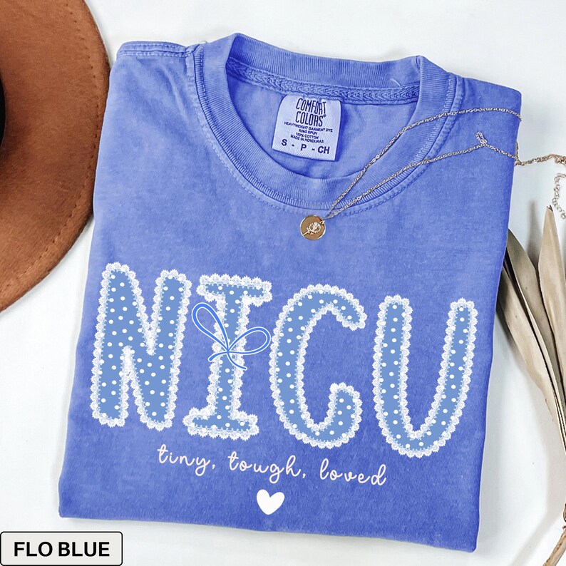 May include: A blue Comfort Colors t-shirt with the word "NICU" in white polka dot letters with a blue bow. The text "tiny, tough, loved" and a white heart are below the word. A gold necklace is draped across the shirt.