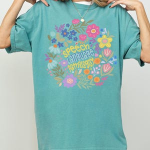 Seafoam Comfort Colors shirt featuring a vibrant circular floral arrangement in pink, yellow, purple, blue, and white. In the center, playful hand-lettered text reads “speech language pathologist” in three stacked lines. “Speech” is in sunny yellow, “Language” is in light blue with playful shading, and “Pathologist” is in orange-yellow with quirky lettering. A tiny heart with a butterfly detail adds charm to the center.