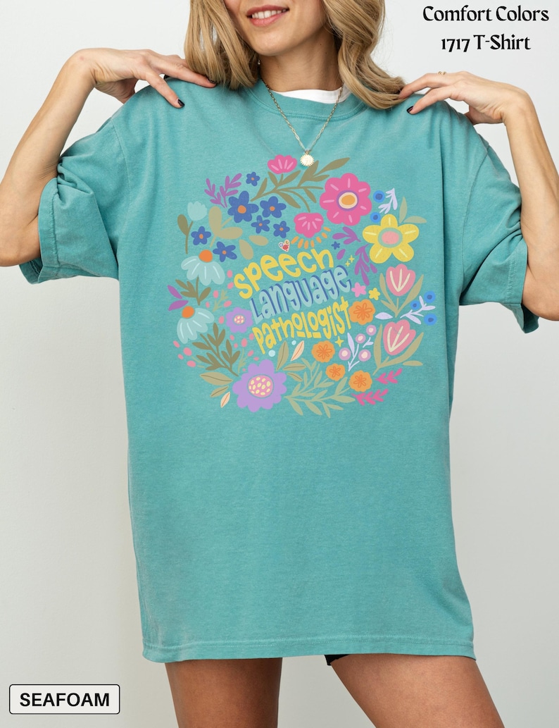 Seafoam Comfort Colors shirt featuring a vibrant circular floral arrangement in pink, yellow, purple, blue, and white. In the center, playful hand-lettered text reads “speech language pathologist” in three stacked lines. “Speech” is in sunny yellow, “Language” is in light blue with playful shading, and “Pathologist” is in orange-yellow with quirky lettering. A tiny heart with a butterfly detail adds charm to the center.