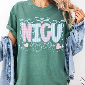 May include: A light green t-shirt with the word "NICU" in pink and blue checkered and striped patterns. The word is surrounded by decorative elements including hearts, stars, and bows.