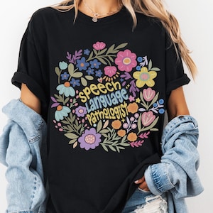 Puede incluir: Camiseta negra con un dise&ntilde;o floral que rodea las palabras "Speech Language Pathologist" en amarillo. El arreglo floral incluye flores rosas, azules, moradas y amarillas. La camiseta es una Comfort Colors 1717 T-Shirt.