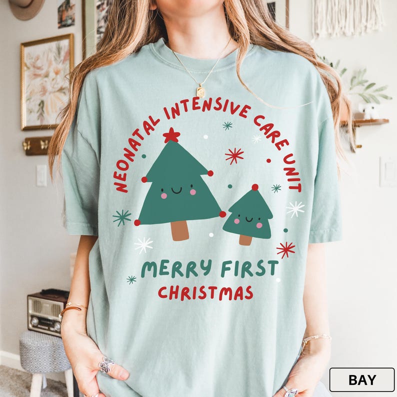 May include: A pale blue t-shirt featuring a festive Christmas design. The text reads "Neonatal Intensive Care Unit" and "Merry First Christmas". The graphic includes two green Christmas trees with red star and dot accents.