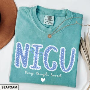 May include: Seafoam green t-shirt with the word "NICU" in blue polka dot letters with a white lace trim. The shirt also has the words "tiny, tough, loved" and a white heart. A gold necklace is draped across the shirt.