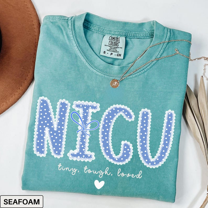 May include: Seafoam green t-shirt with the word "NICU" in blue polka dot letters with a white lace trim. The shirt also has the words "tiny, tough, loved" and a white heart. A gold necklace is draped across the shirt.