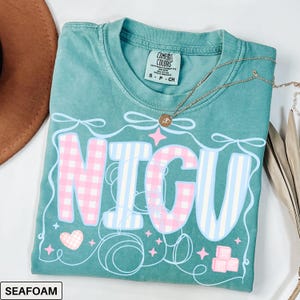 A folded seafoam Comfort Colors T-shirt with "NICU" spelled out in large, patchwork-style letters. The "N" and "C" are soft pink gingham, while the "I" and "U" are light blue and white vertical stripes. Surrounding the letters are pastel pink ribbons, looping thread, sparkles, a pink gingham heart, and pink ABC building blocks. The style is sweet, nurturing, and whimsical, perfect for NICU nurses and staff.