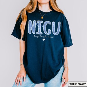 May include: Navy blue t-shirt with the word "NICU" in light blue, polka-dotted letters. Below the word are the words "tiny, tough, loved" and a small white heart. The shirt is a comfortable, casual style.