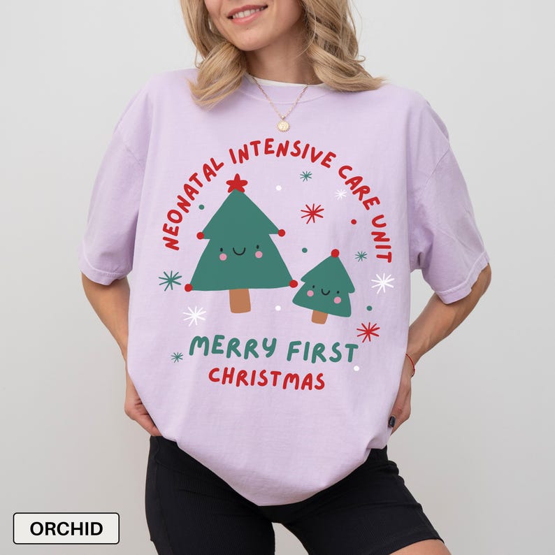 May include: Lilac t-shirt with a Christmas-themed design. The shirt displays a green Christmas tree graphic and the text "Neonatal Intensive Care Unit Merry First Christmas" in red and green. The t-shirt has a loose fit.