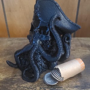 Puede incluir: Un cráneo de pirata negro impreso en 3D con una barba de pulpo que lleva un sombrero de pirata negro. El cráneo está sentado sobre una superficie de madera con un rollo de monedas al lado.