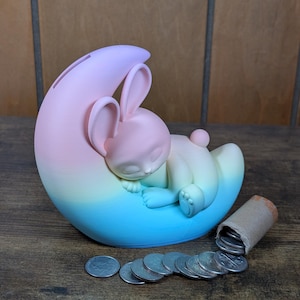 May include: A pastel-colored bunny-shaped piggy bank, sleeping on a crescent moon. The bank is a gradient of pink, yellow, and blue. A roll of coins and loose change are next to the bank.