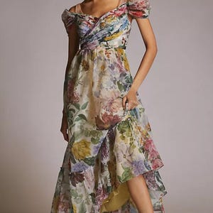 May include: A floral print dress with a layered skirt and off-the-shoulder sleeves. The dress features a mix of colorful flowers on a light background. The model is wearing nude-colored sandals.