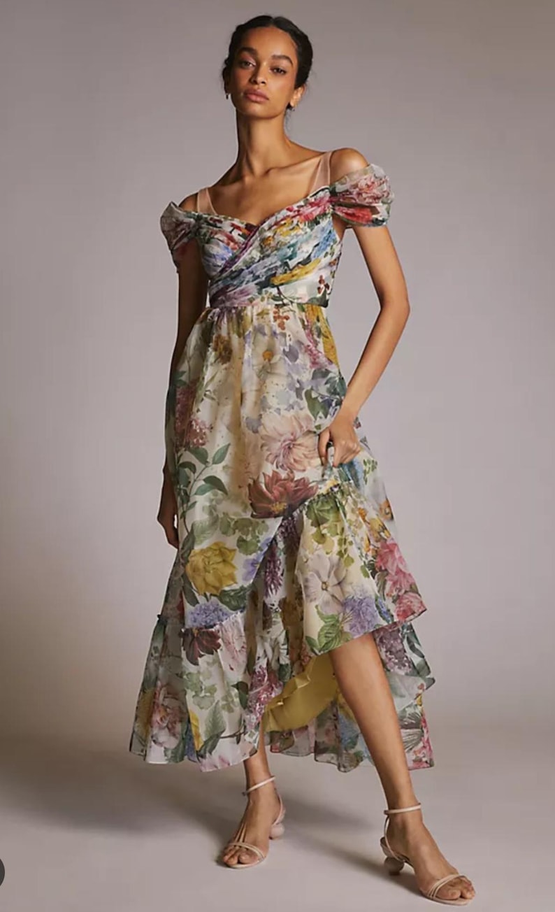 May include: A floral print dress with a layered skirt and off-the-shoulder sleeves. The dress features a mix of colorful flowers on a light background. The model is wearing nude-colored sandals.