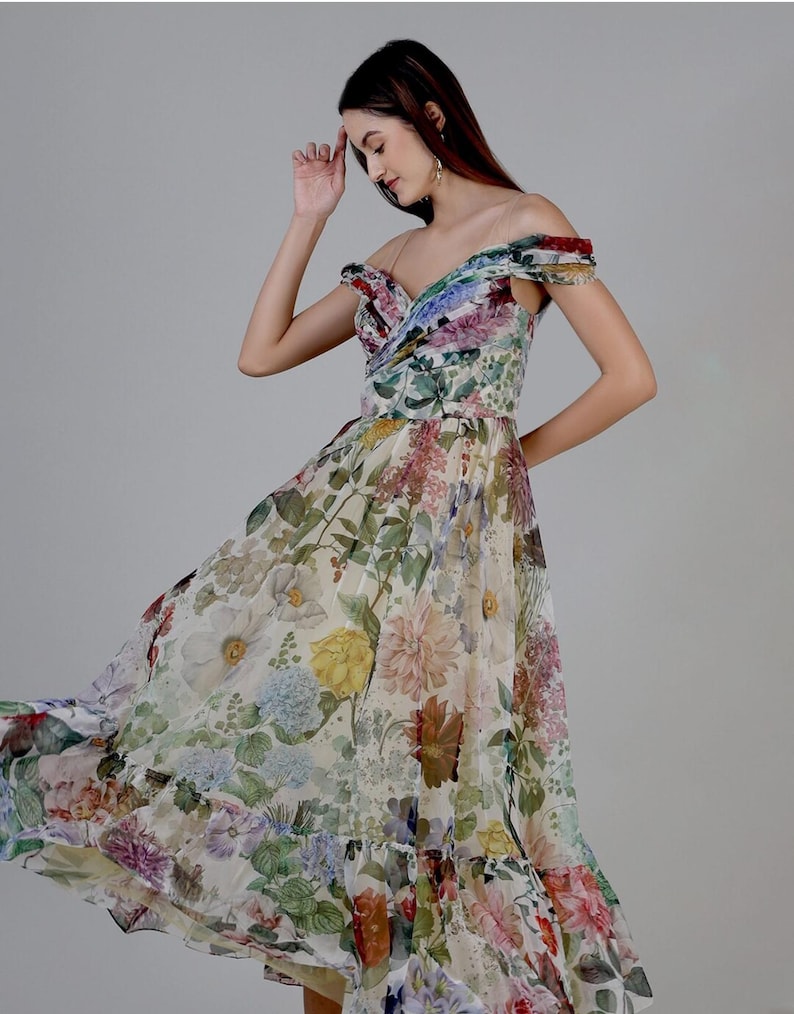 May include: Off-the-shoulder floral print dress with a flowing skirt. The dress features a variety of colorful flowers in shades of pink, purple, yellow, and green against a cream background. The dress has a fitted bodice and a ruffled hemline.