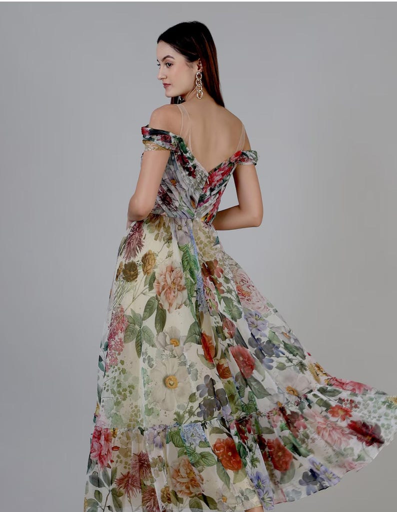 May include: A floral print off-the-shoulder dress with a flowing skirt. The dress features a variety of flowers in shades of red, pink, yellow, and green on a white background. The dress has a low back and sheer straps.