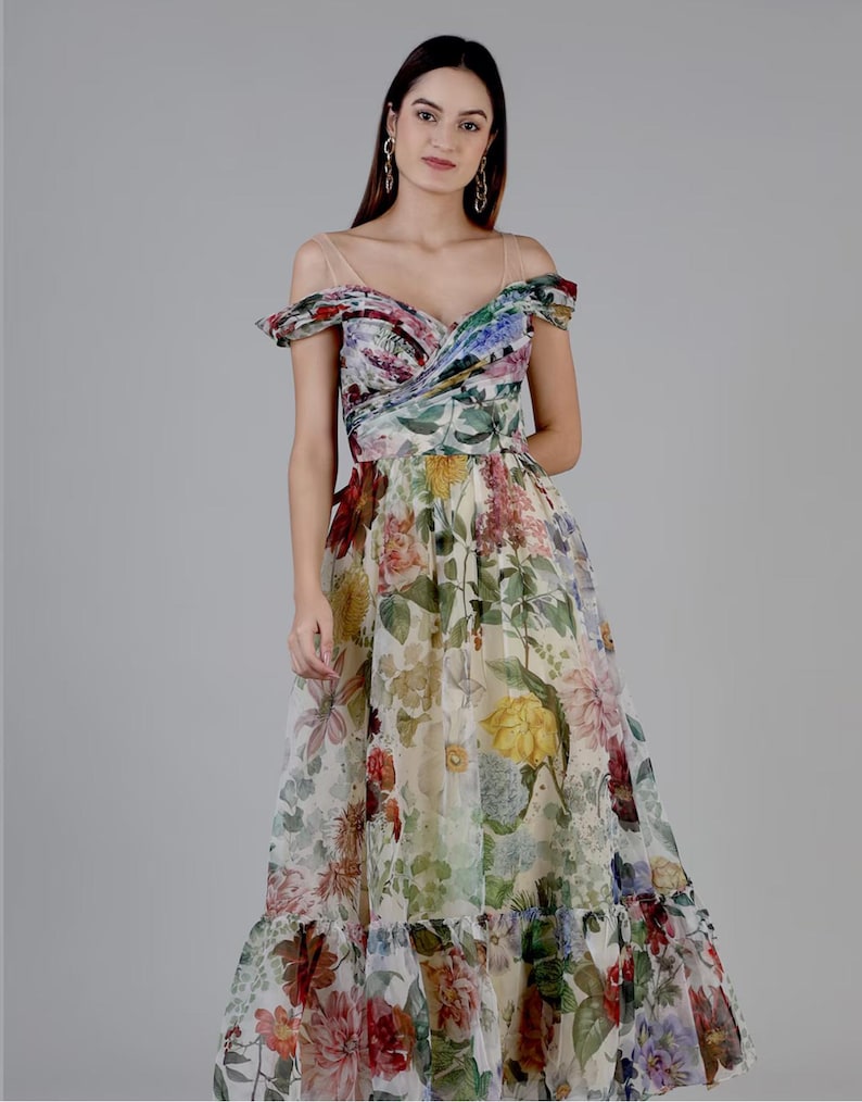 May include: A floral print maxi dress with an off-the-shoulder neckline. The dress features a variety of colorful flowers, including red, yellow, and purple blooms, set against a cream background. The dress has a flowing skirt and a ruffled hem.