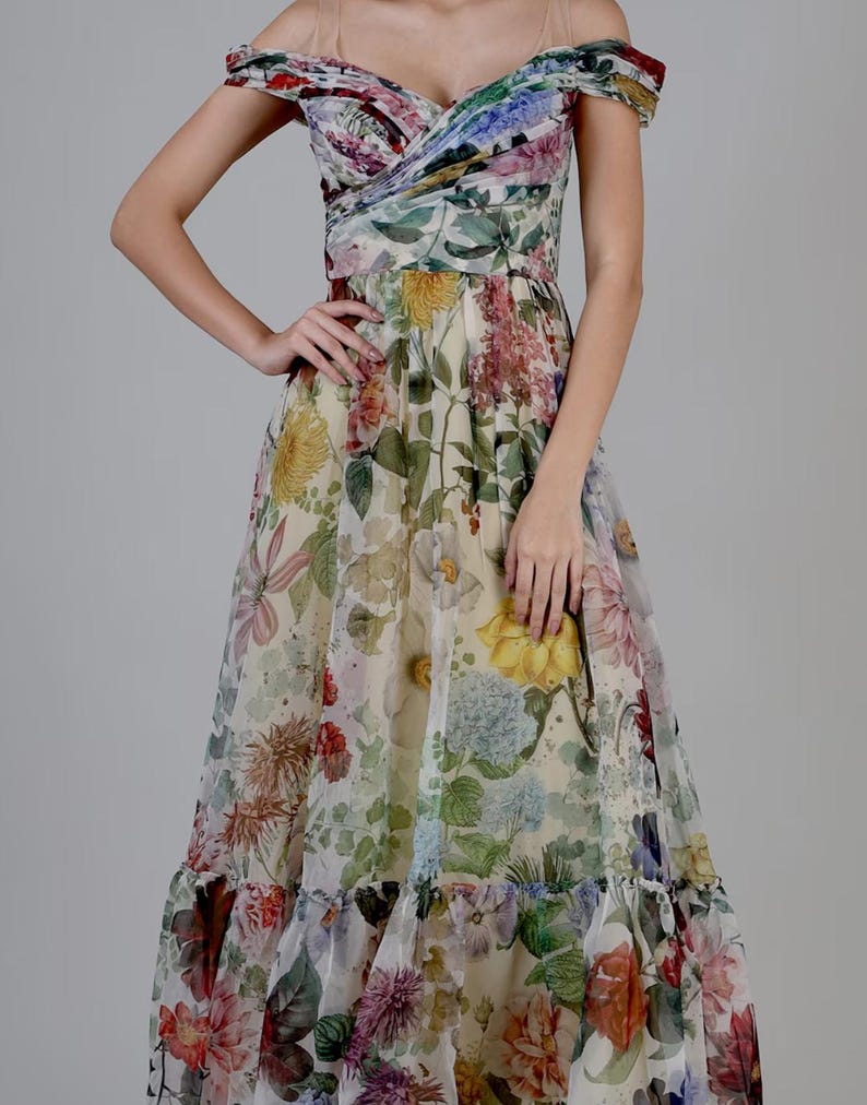 May include: A floral print off-the-shoulder dress with a sweetheart neckline. The dress features a variety of colorful flowers, including yellow, pink, and blue, on a cream-colored background. The dress has a flowing skirt and a ruffled hem.