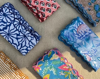 Pode incluir: Seis guardanapos de tecido dobrados com bordas onduladas. Os guardanapos apresentam vários padrões, incluindo florais e listrados, em tons de rosa, azul, castanho e amarelo. Estão dispostos sobre uma superfície de cor neutra.