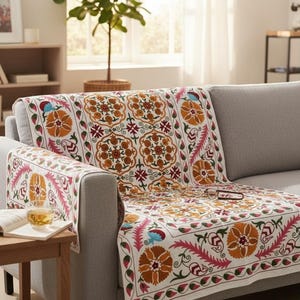 May include: A white sofa cover with a vibrant floral pattern in orange, pink, and blue. The cover is draped over a gray sofa in a well-lit living room. A small wooden side table with a cup of tea and a book is in the foreground.