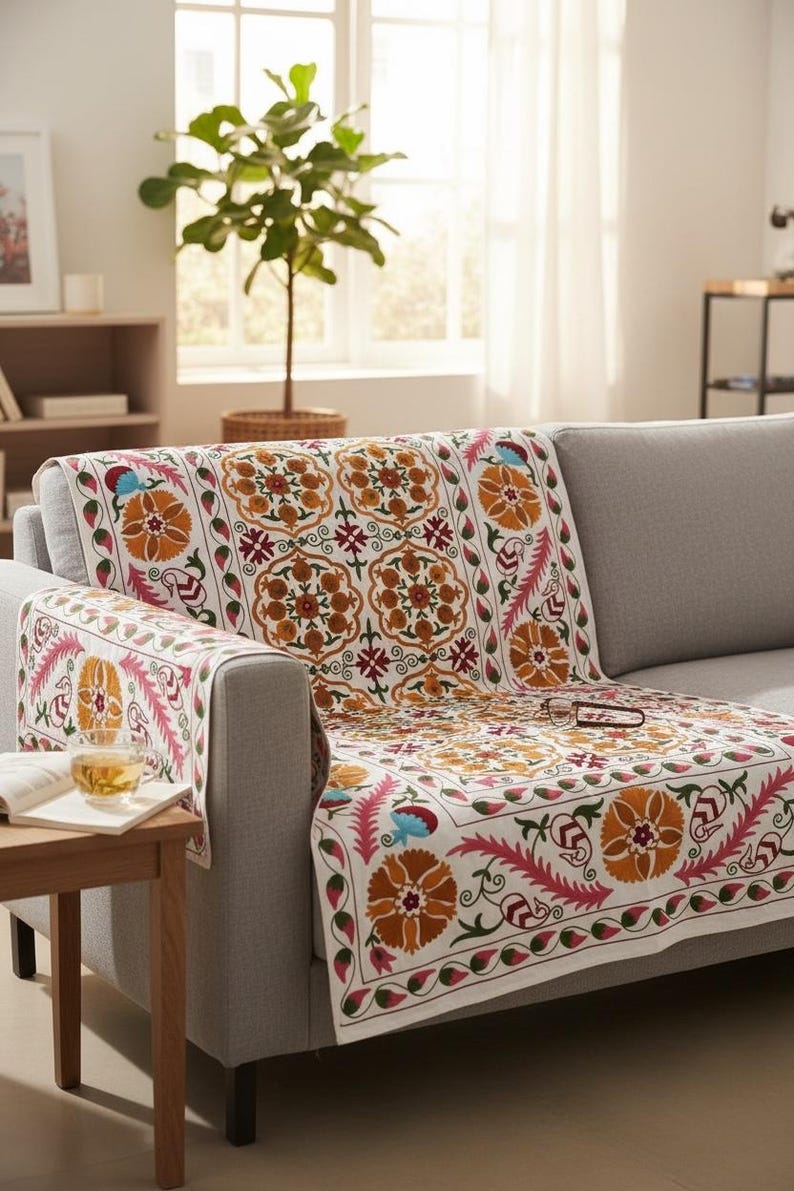 May include: A white sofa cover with a vibrant floral pattern in orange, pink, and blue. The cover is draped over a gray sofa in a well-lit living room. A small wooden side table with a cup of tea and a book is in the foreground.