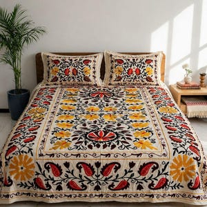 Puede incluir: Juego de cama bordado crema con dos fundas de almohada. El diseño floral presenta detalles en amarillo, rojo y marrón. La colcha está sobre un marco de cama de madera, con una planta en el fondo.