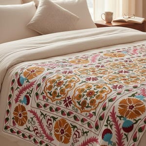 May include: A cream-colored bedspread with a detailed floral pattern in shades of orange, pink, blue, and green. The bed is made with cream-colored pillows and a matching comforter. The bed is in a well-lit room.