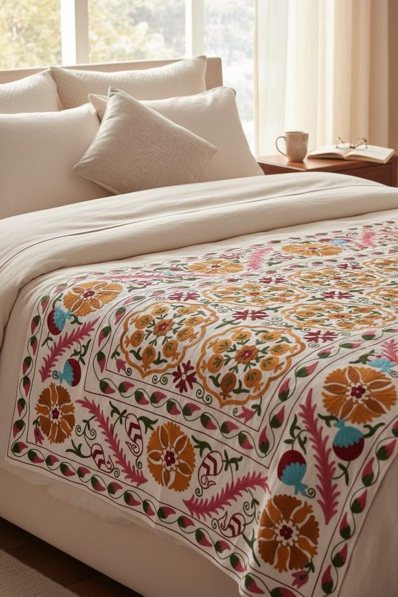 May include: A cream-colored bedspread with a detailed floral pattern in shades of orange, pink, blue, and green. The bed is made with cream-colored pillows and a matching comforter. The bed is in a well-lit room.
