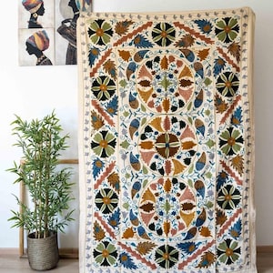 May include: Cream-coloured textile featuring a large, intricate floral design in shades of green, blue, brown, and yellow. The textile is rectangular, possibly a wall hanging or tapestry. Decorative art pieces are visible in the background.