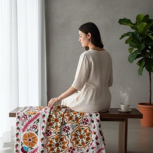 May include: A white textile with a colorful floral pattern, featuring orange, pink, and green accents, is draped over a dark wooden bench. The textile is the focal point, showcasing intricate embroidery. A woman sits on the bench near a cup of tea.