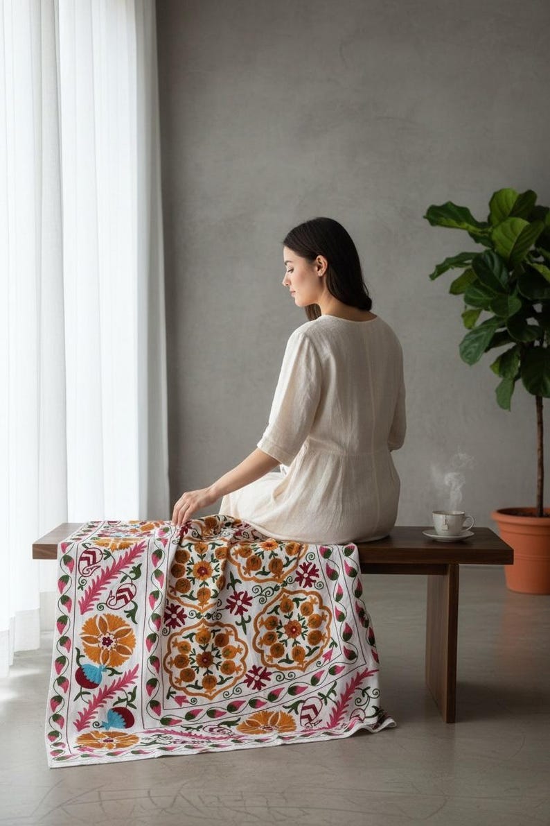 May include: A white textile with a colorful floral pattern, featuring orange, pink, and green accents, is draped over a dark wooden bench. The textile is the focal point, showcasing intricate embroidery. A woman sits on the bench near a cup of tea.
