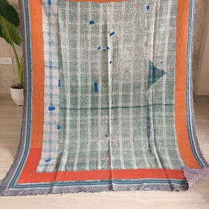 Peut inclure: Un jeté de lit rectangulaire présentant un motif central à carreaux verts et blancs, bordé d'une large bande orange et d'un fin liseré bleu rayé. Le tissu présente des coutures visibles et de petites pièces bleues éparpillées sur la surface.