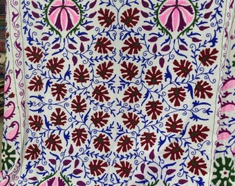 May include: White textile with detailed embroidery. The design includes pink, burgundy, blue, and green floral patterns. Large pink flower-like shapes are centered, surrounded by leaf and vine motifs. The textile is likely a decorative item.
