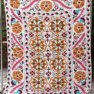 May include: White textile with intricate embroidery featuring floral and geometric patterns in shades of orange, pink, red, blue, and green. The design includes large floral motifs and decorative borders, creating a vibrant and detailed textile.