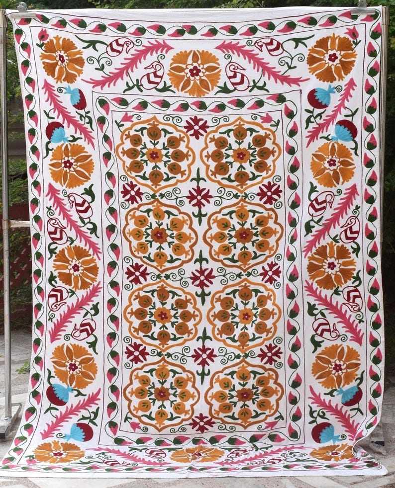 May include: White textile with intricate embroidery featuring floral and geometric patterns in shades of orange, pink, red, blue, and green. The design includes large floral motifs and decorative borders, creating a vibrant and detailed textile.