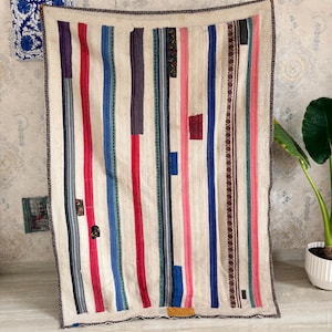 May include: A rectangular textile throw featuring a cream-colored base with vertical stripes in various widths and colors, including red, blue, pink, purple, and brown. The fabric displays a textured weave with visible stitching and patchwork details.