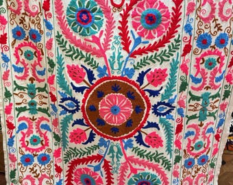May include: White textile with vibrant floral embroidery in red, pink, blue, and green. The design features a central medallion and repeating floral patterns along the borders. The embroidery is detailed and colourful, creating a decorative textile.