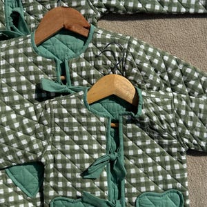 Può includere: Giacche trapuntate a quadri verdi e bianchi con tasche a forma di cuore e chiusure con fiocco. Le giacche sono esposte su grucce di legno, mostrando il loro design e la loro consistenza. Realizzate in un tessuto trapuntato morbido.