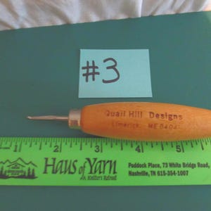 May include: A wooden-handled seam ripper with a metal tip, positioned above a green ruler showing a length of approximately 6 inches. The handle is printed with the text Quail Hill Designs Limerick ME 04048. A light blue note with #3 is visible.