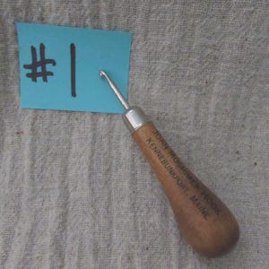 May include: A rug hooking tool with a metal hook and a wooden handle. The handle is printed with the text JOAN MOSHIMER HOOK KENNEBUNKPORT, MAINE. A light blue rectangular paper tag with the handwritten text #1 is positioned next to the tool.