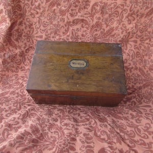Peut inclure: Une boîte en bois rectangulaire avec un couvercle à charnière. La boîte est de couleur marron moyen avec une petite étiquette métallique qui indique "Workbox". La boîte est posée sur un tissu à motifs.