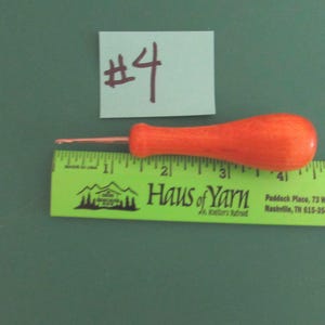 May include: A crochet hook with a bright orange wooden handle positioned above a green ruler. The hook measures approximately 3.5 inches in total length. A small light blue paper tag with the handwritten text #4 is placed above the hook.