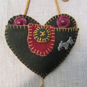 May include: Handmade heart-shaped felt ornament with a dark green base, red accents, and gold stitching. Features two purple buttons, a small metal dog charm, and a gold cord for hanging. A unique, handcrafted item.